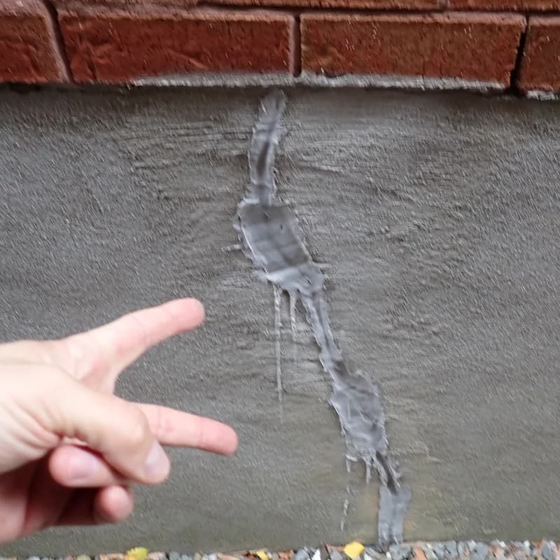 Main qui pointe une trace d'infiltration d'eau sur une fondation en crépi, sous un mur de brique
